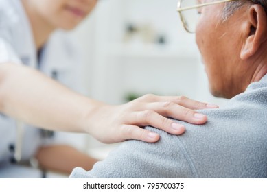 Young Asian Female Doctor Putting Her Hand On Senior Male Patient Shoulder For Comforting In Medical Room At The Hospital. Diseases, Ill, Symptom, Health Care Talking Of Elderly.