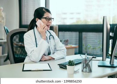 young asian female doctor using mobile phone talking while sitting in hospital office. happy woman medical worker having joyful conversation with husband in break time. nurse resting in clinic. - Powered by Shutterstock