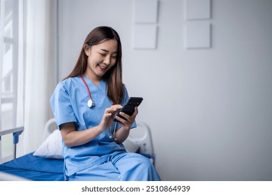 Young asian female doctor wearing blue scrubs is sitting on a hospital bed and using a smartphone - Powered by Shutterstock