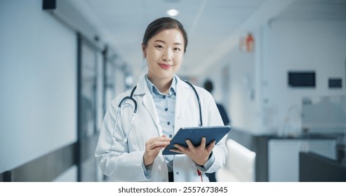 Young Asian Female Doctor in White Coat, with a Stethoscope Around Her Neck, Standing in a Bright Hospital Hallway and Using Tablet. Professional Medical Worker Looks at Camera with a Smile - Powered by Shutterstock