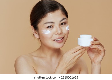 Young Asian Female With Facial Cream Jar And Eye Cream On Face Against Beige Background.