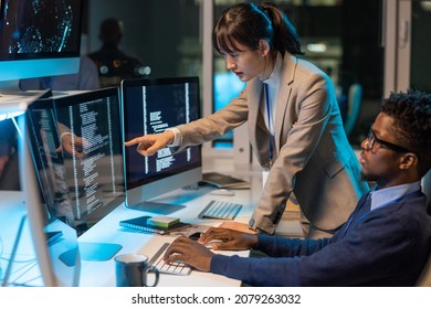 Young Asian female it-engineer pointing at data on computer screen while discussing working point with African male colleague