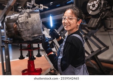 Young asian female mechanic inspecting a gearbox illuminated with a flashlight in a car repair shop - Powered by Shutterstock