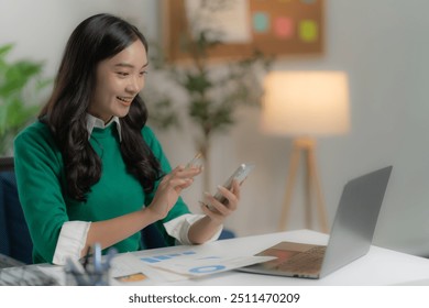 Young Asian female office worker wearing green shirt working in office using laptop and mobile phone. - Powered by Shutterstock