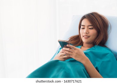 Young Asian Female Patient Holding And Checking Mobile Phone While Laying On The Bed In A Hospital Room. Health Care And Medical Insurance Concept.