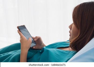 Young Asian Female Patient Holding And Checking Mobile Phone While Laying On The Bed In A Hospital Room. Health Care And Medical Insurance Concept.