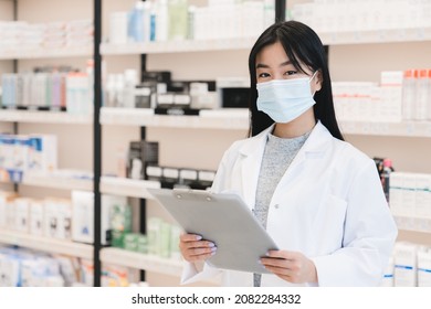 Young asian female pharmacist druggist in white coat and protective medical mask against coronavirus holding clipboard while looking at camera in drugstore pharmacy