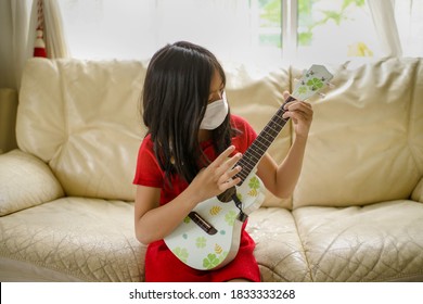 Young Asian Female Playing Ukelele At Home During Pandemic While Wearing Mask.Girl Child With String Instrument.Acoustic Music.Homeschooling And Leisure.