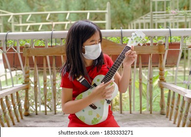 Young Asian Female Playing Ukelele At Home During Pandemic While Wearing Mask.Girl Child With String Instrument.Acoustic Music.Homeschooling And Leisure.