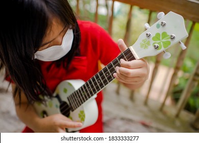 Young Asian Female Playing Ukelele At Home During Pandemic While Wearing Mask.Girl Child With String Instrument.Acoustic Music.Homeschooling And Leisure.