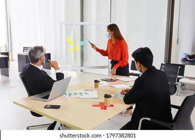 Young Asian Female Presenting Sales Graph On The Board To His Senior Male Manager And His Colleagues. All Wearing Masks And Keep Distance For Good Health And Hygiene At Workplace  During Covid 19