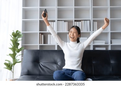 Young Asian female reading good news on phone. Surprised asian woman celebrating victory on smartphone on sofa at home. - Shutterstock ID 2287363335