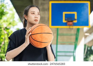 Young asian female in sportswear holding ball and looking away while playing basketball on playground with green trees in background - Powered by Shutterstock