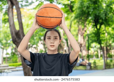 Young asian female in sportswear holding ball and looking away while playing basketball on playground with green trees in background - Powered by Shutterstock