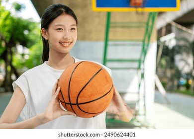 Young asian female in sportswear holding ball and looking away while playing basketball on playground with green trees in background - Powered by Shutterstock