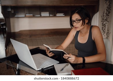 Young asian female student in glasses using laptop, read a book. Cozy office workplace, remote work, E learning concept. - Powered by Shutterstock