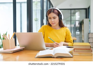 Young asian female student in glasses studying learning online coureses and writing down notes while sitting at desk and using laptop and textbooks wearing headphone. Online study and learning concept