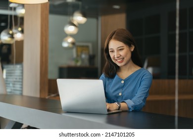 Young Asian Female Student Sitting Taking Notes And Using Laptop Computer At Table.