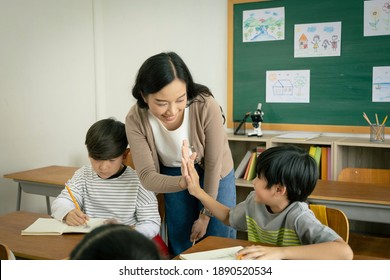 A Young Asian Female Teacher And An Elementary School Boy Clapping Together Hands In The Classroom. The Teacher Congratulates The Students On The Success