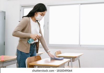 Young Asian Female Teacher In A Medical Mask Cleans Up The Desk In The Classroom Before Students Return To School After Covid-19 Quarantine And Lockdown. Healthcare Of Pupils And Students Concept.