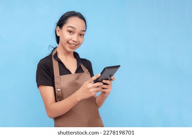 A young asian female waitress or barista taking an order on her tablet while smiling. Wearing a brown apron and black shirt welcoming a customer and against a light blue background. - Shutterstock ID 2278198701