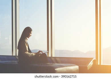 Young Asian Female Is Watching News On Digital Tablet, While Is Sitting In Airport Interior Against Window With Copy Space Area.Attractive Chinese Woman Is Using Portable Computer And Waiting Her Fly 