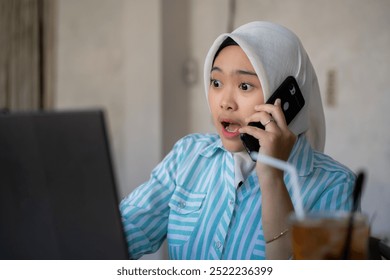 a young Asian female worker wearing a hijab contacting her coworkers via voice call while working remotely at a cafe, the concept of remote work, working flexibly from anywhere. - Powered by Shutterstock