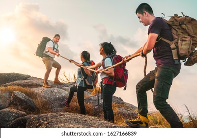 Young Asian Four Hikers Climbing Up On The Peak Of Mountain. People Helping Each Other Hike Up A Mountain At Sunlight. Giving A Helping Hand. Climbing. Helps And Team Work Concept