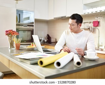 Young Asian Free Lance Designer Sitting At Kitchen Counter Creating A Design Using Pen Tablet And Laptop Computer.