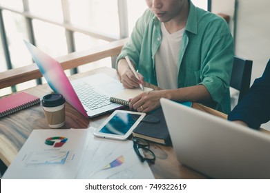 Young Asian Freelance Worker Writing On Small Notebook While Working With Colleague In Coffee Shop. Freelancer On Workday Activity In Coworking Space With Photo Filter Effect