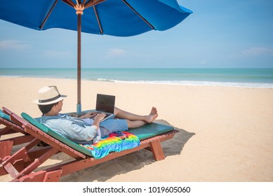 Young Asian Freelancer Man Lying On Resort Beach Bench With Blue Umbrella Overhead Working With Laptop Computer. Digital Nomad Life Or Freelance Lifestyle. Remote Work On Summer Vacation.