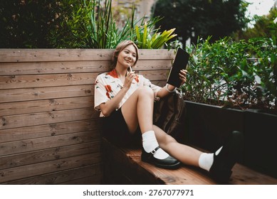 Young Asian freelancer smiling while working on tablet with stylus, relaxed in cozy wooden nook filled with plants, enjoying digital creativity and comfortable mobile workflow.