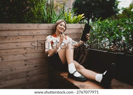 Young Asian freelancer smiling while working on tablet with stylus, relaxed in cozy wooden nook filled with plants, enjoying digital creativity and comfortable mobile workflow.