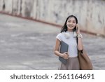 Young asian freelancer woman is walking outdoors while talking on the phone, holding a laptop under her arm and looking away, with wireless headphones around her neck