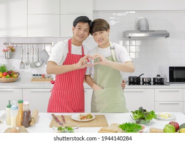 Young Asian Gay Couple Wearing Apron, Looking Camera And Happy Making Heart Shape With Hands Together During Cooking In The Kitchen.