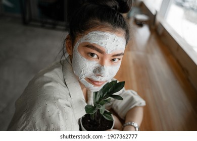 Young Asian Girl With A Clay Mask On Her Face Hugs A Flower Pot, The Concept Of Natural Products Lying In The Base Of The Mask