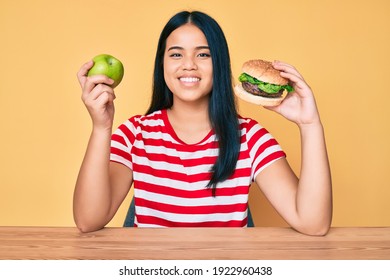 Young Asian Girl Comparing Burger And Healthy Apple Smiling With A Happy And Cool Smile On Face. Showing Teeth. 