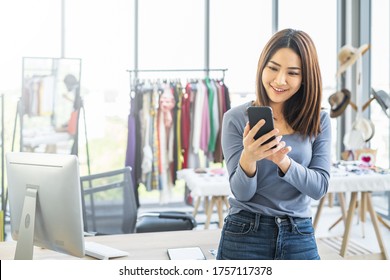 Young Asian Girl Fashion Shop Owner Smiling Looking At Mobile Phone Device Looking Happy Inside A Clothing Store.