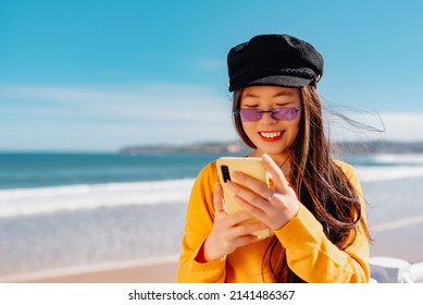 Young Asian Girl With Glasses And Beret Consulting Maps On Her Smart Phone While Sightseeing In A European City. Chinese Woman Using A Smartphone