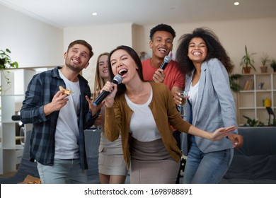 Young Asian Girl Holding Microphone And Singing While Playing Karaoke With Best Friends At Home. Group Of People Having Fun Together, Drinking And Eating Pizza. Karaoke Party