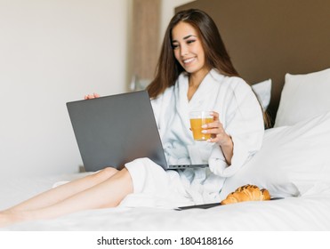 Young Asian Girl With Long Hair In White Robe Using Laptop Having Breakfast Croissant And Orange Juice In Bed Of Hotel Room