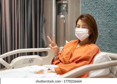 
Young Asian Girl  Lying In The Hospital Bed, Wear A Mask And Lift 2 Inches Symbol To Indicate That It's Still Fine