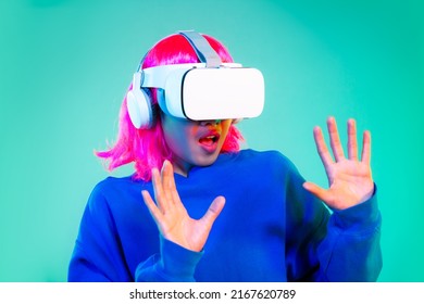 Young Asian Girl Pink Color Hairstyle In Blue Sweatshirt Wearing White Vr Headset Watching Playing And Exciting On The Green Screen Background. Cyborg And Metaverse Concept.