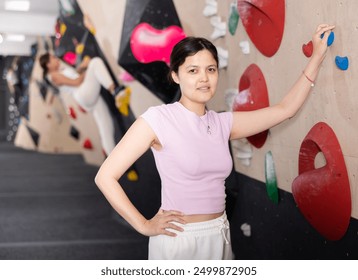 Young Asian girl poses near steep climbing wall in gym and leans against hook. Woman is resting after workout and leaning against artificial wall for sports activities - Powered by Shutterstock