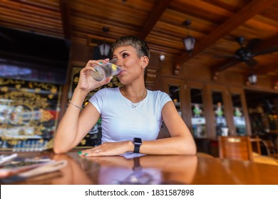 Ein junges asiatisches Mädchen sitzt allein an einem Holztisch in einem Restaurant und trinkt ein Glas Wasser. Porträt einer ziemlich lächelnden malaysischen Frau, sitzend an einem Tisch in einem Café in Kuala Lumpur, Malaysia. 
