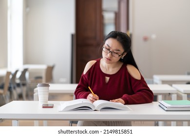 Young Asian Girl Student Making Assessments From Book For Study. Female Preparing For Exam In University, College Or High School, Make Homework Sitting In Empty Classroom Or Library. Education Concept