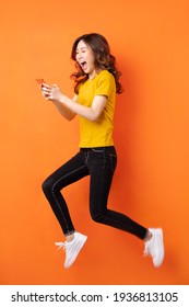 Young Asian Girl Using The Phone And Jumping On Orange Background