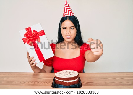 Young asian girl wearing birthday hat holding present annoyed and frustrated shouting with anger, yelling crazy with anger and hand raised 