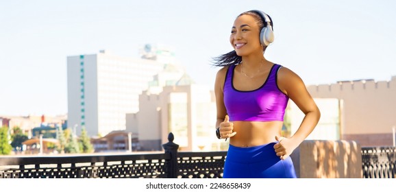 Young Asian girl in white headphones runs early in the morning against the backdrop of the cityscape. Attractive looking woman keeping fit and healthy. Forrmat 20x9.