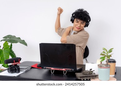 A young Asian graphic designer stretching his stiff shoulder while working at his desk with headphones and a laptop. - Powered by Shutterstock